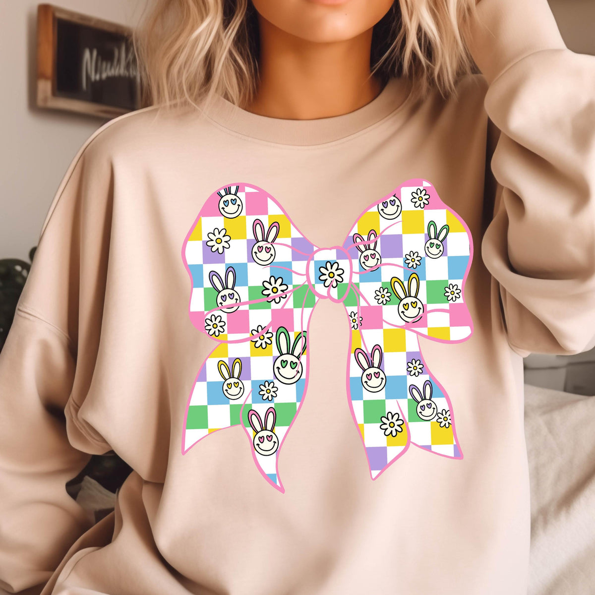 May include: A pink sweatshirt with a large, colorful bow printed on the front. The bow is made up of checkered squares with smiling bunny faces and flowers. 