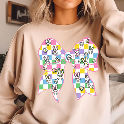 May include: A pink sweatshirt with a large, colorful bow printed on the front. The bow is made up of checkered squares with smiling bunny faces and flowers. 