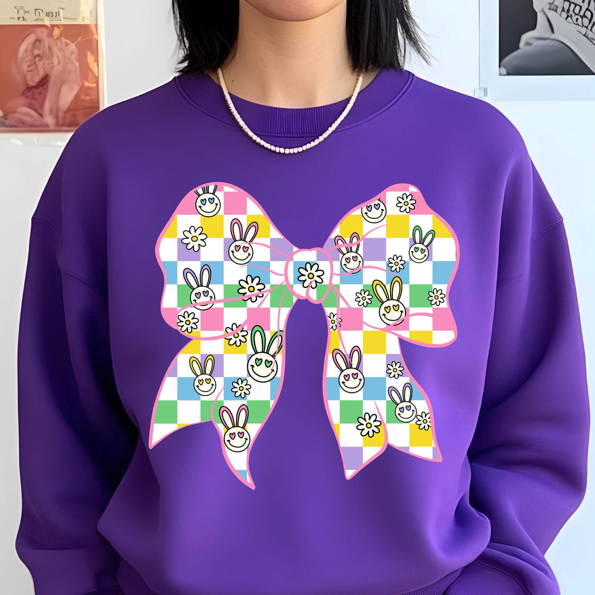 May include: Purple sweatshirt with a large pink bow printed on the front. The bow is checkered with pastel colors and has smiling bunny faces and flowers.