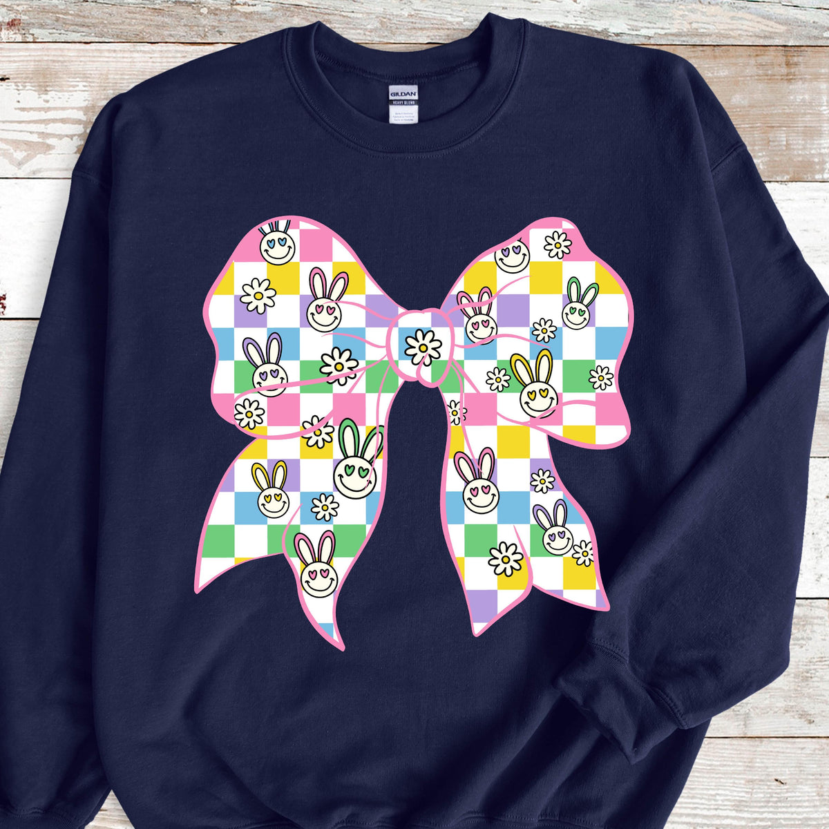 May include: A navy blue sweatshirt with a pink bow. The bow is patterned with a checkered design of pastel colors, including yellow, green, purple, and pink. The pattern includes bunny faces with hearts for eyes and white flowers.