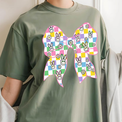 May include: A green t-shirt with a pink bow printed on the front. The bow is checkered with pink, yellow, green, and blue squares. Each square has a small white flower and a cartoon bunny with a smiley face.