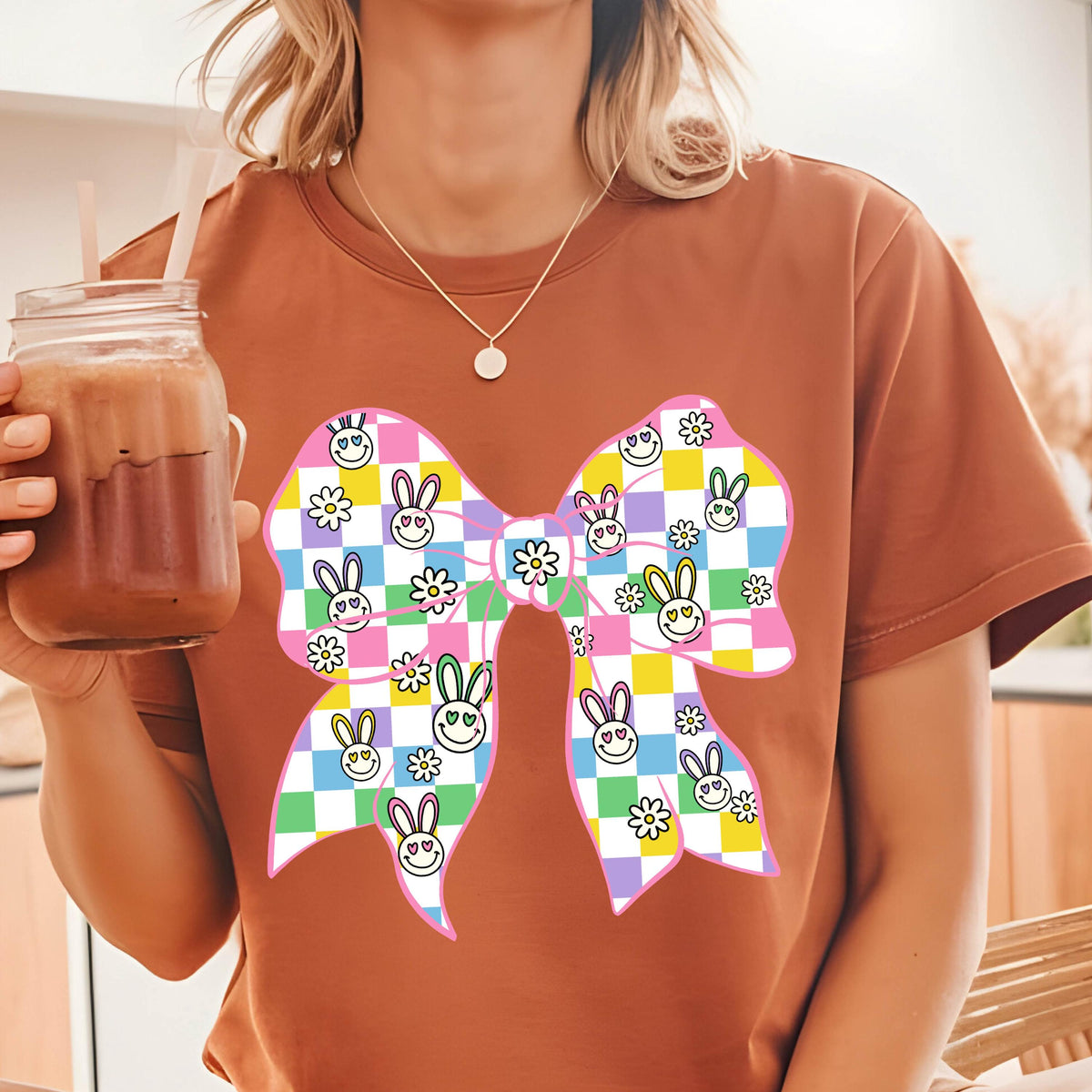 May include: A brown t-shirt with a large pink, yellow, green, and blue bow printed on the front. The bow is made up of checkered squares with bunny faces and flowers.