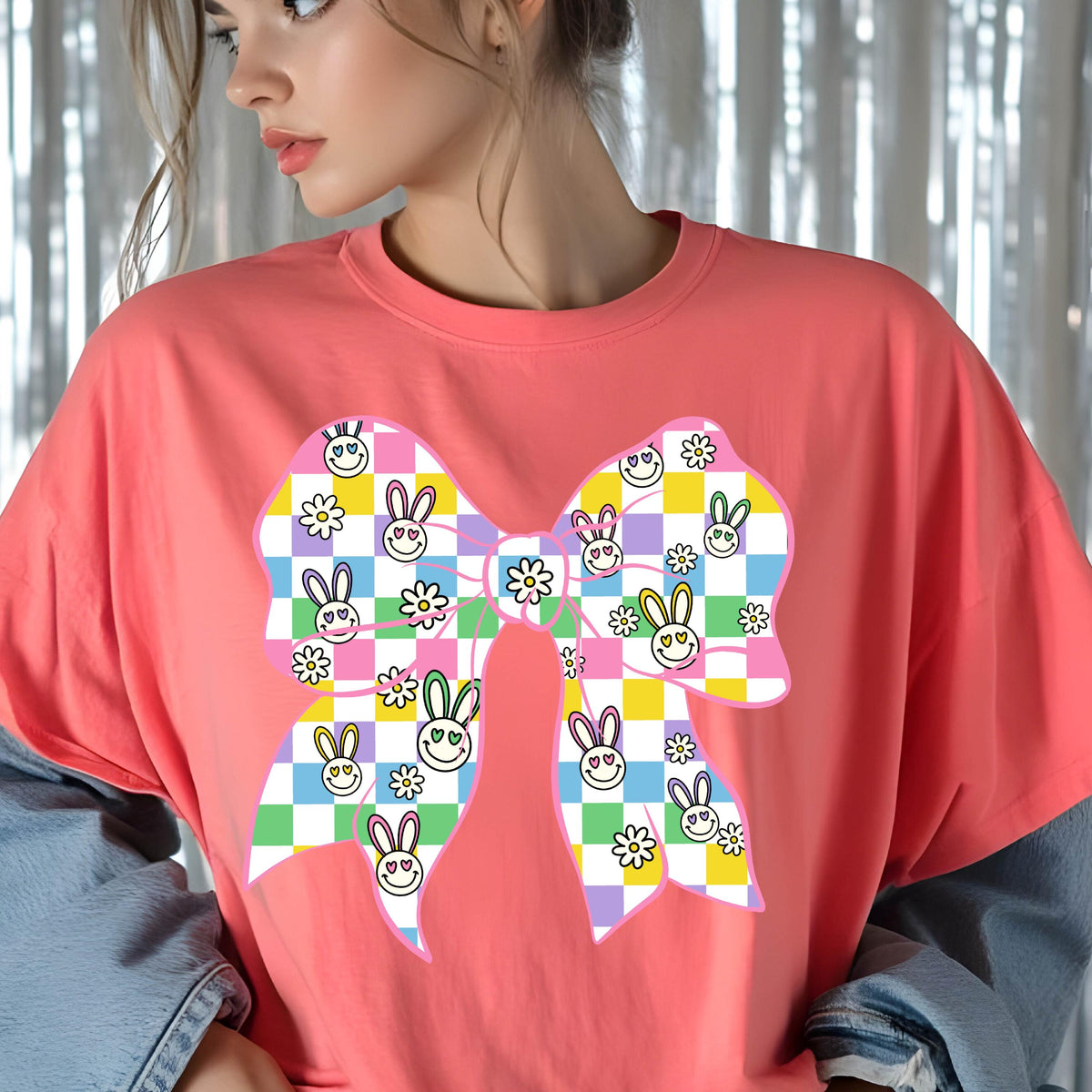 May include: A pink t-shirt with a large, colorful bow design. The bow is made up of checkered squares in shades of pink, yellow, green, and blue. The squares are decorated with cartoon bunny faces and daisies.