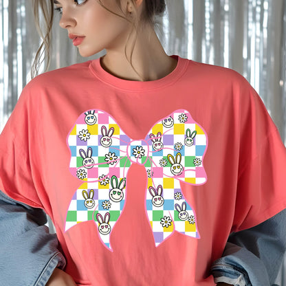 May include: A pink t-shirt with a large, colorful bow design. The bow is made up of checkered squares in shades of pink, yellow, green, and blue. The squares are decorated with cartoon bunny faces and daisies.