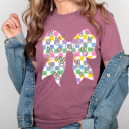 May include: A pink t-shirt with a large, colorful bow printed on the front. The bow is made up of checkered squares in shades of pink, blue, green, and yellow, with a pattern of smiling bunnies and flowers.