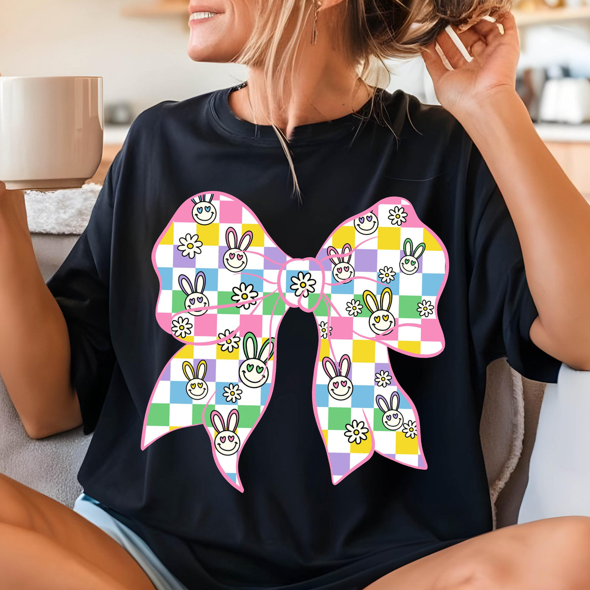 May include: A black t-shirt with a large pink bow printed on the front. The bow is made up of checkered squares in pink, yellow, green, and purple. Each square has a small white flower and a bunny face with a smiley face.