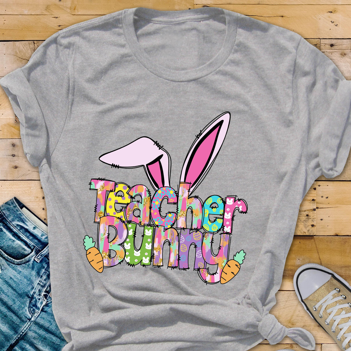 May include: A gray t-shirt featuring a colorful "Teacher Bunny" graphic with bunny ears and carrots. The text is in a playful font with a mix of patterns and colors, perfect for Easter or a teacher's gift.