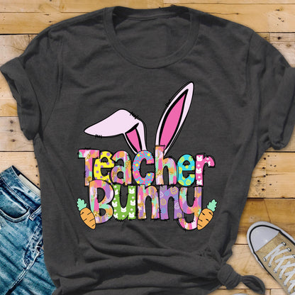 May include: Dark gray t-shirt featuring a colorful graphic. The graphic includes bunny ears, the words "Teacher Bunny" in a patterned font, and two carrot illustrations. The shirt is a casual style.