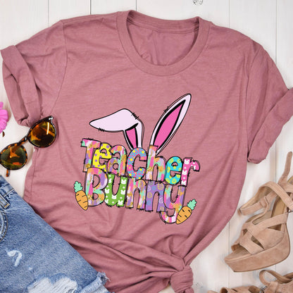 May include: A dusty rose-colored t-shirt with the words "Teacher Bunny" in colorful, patterned letters. The design includes bunny ears and carrots, perfect for Easter or a teacher's gift.