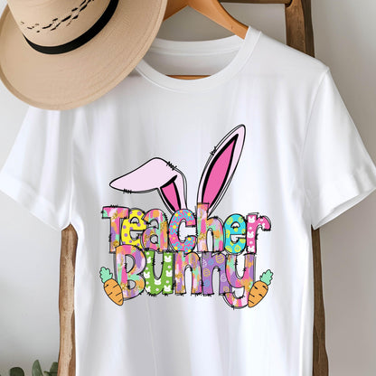May include: White t-shirt featuring the words "Teacher Bunny" in colorful, patterned letters, with bunny ears and carrots. The design is perfect for Easter or a teacher's gift.
