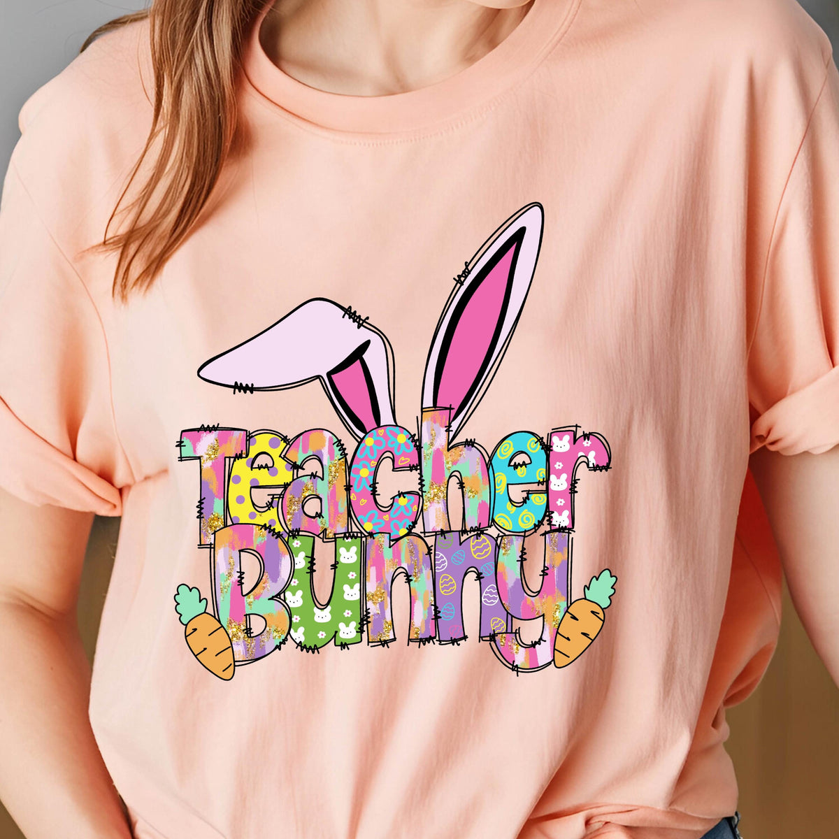 May include: Peach-colored t-shirt featuring the words "Teacher Bunny" in a colorful, patterned font. The design includes bunny ears and carrots, perfect for Easter or a teacher's gift.