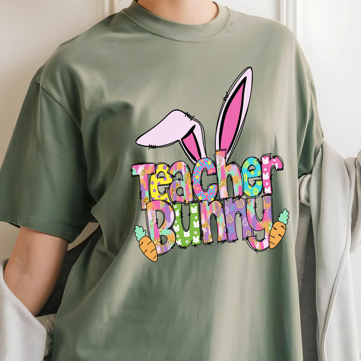 May include: Sage green t-shirt with a colorful "Teacher Bunny" graphic. The design features bunny ears, carrots, and the words in a playful font. A fun Easter-themed tee for teachers.