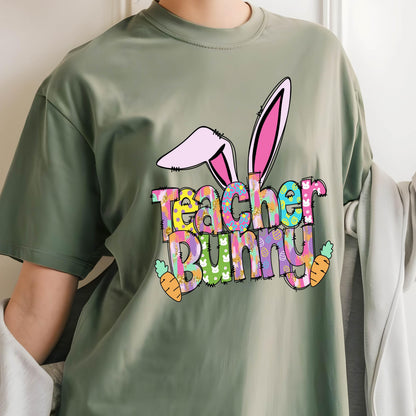 May include: Sage green t-shirt with a colorful "Teacher Bunny" graphic. The design features bunny ears, carrots, and the words in a playful font. A fun Easter-themed tee for teachers.