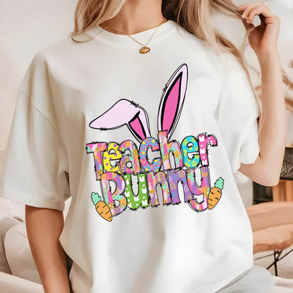 May include: White t-shirt with the words "Teacher Bunny" in colorful, patterned letters. The design includes bunny ears and carrots, perfect for Easter or a teacher's gift.