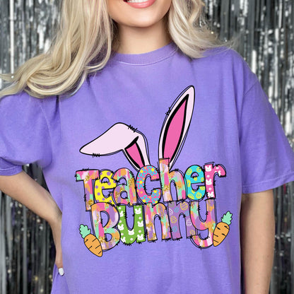 May include: A purple t-shirt with a colorful graphic that says "Teacher Bunny." The design includes bunny ears and two orange carrots. The text is in a playful, multi-colored font, perfect for Easter or a teacher's gift.