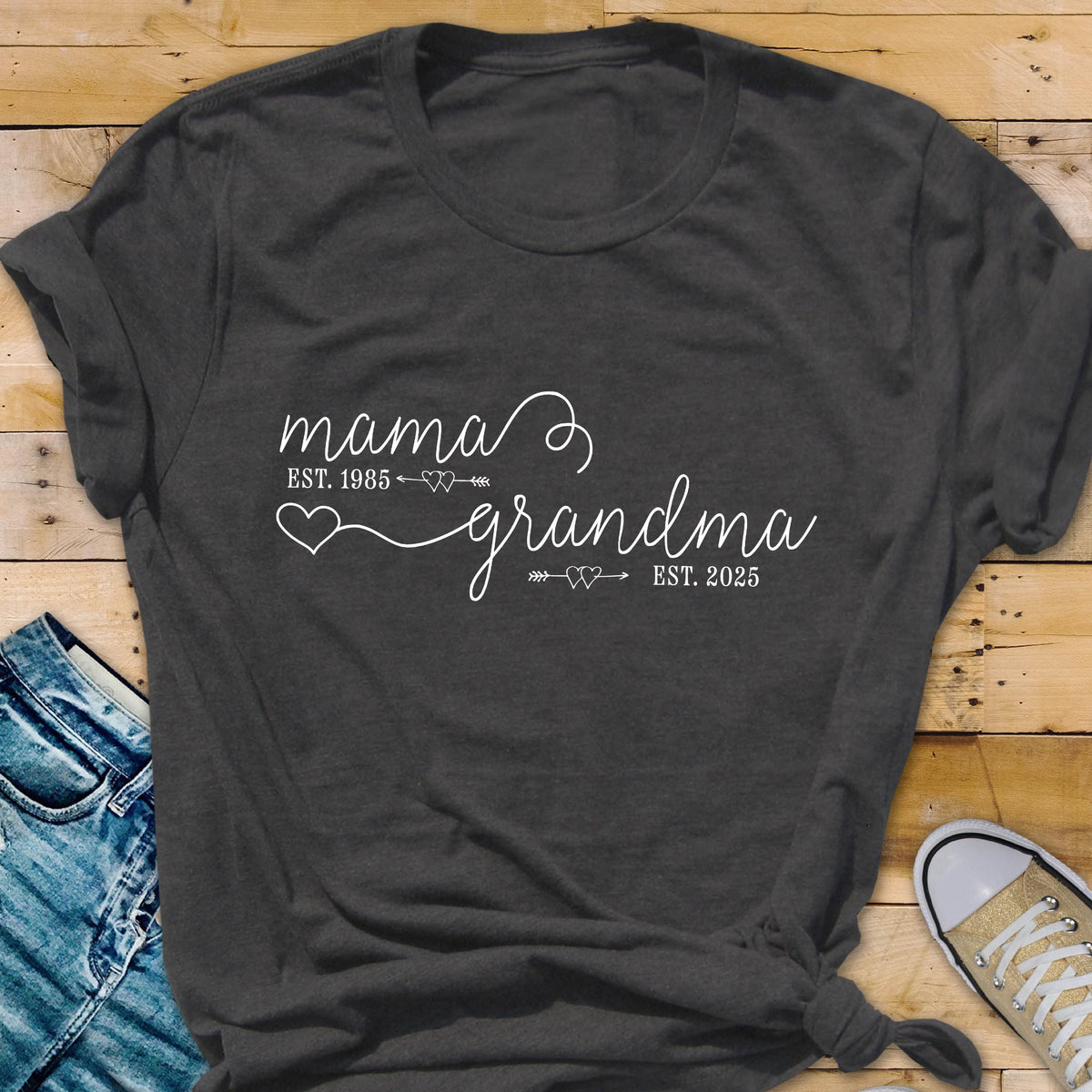 May include: A dark gray t-shirt with white text that reads "mama est. 1985 grandma est. 2025". The text is in a cursive font and includes a heart and arrow graphic.