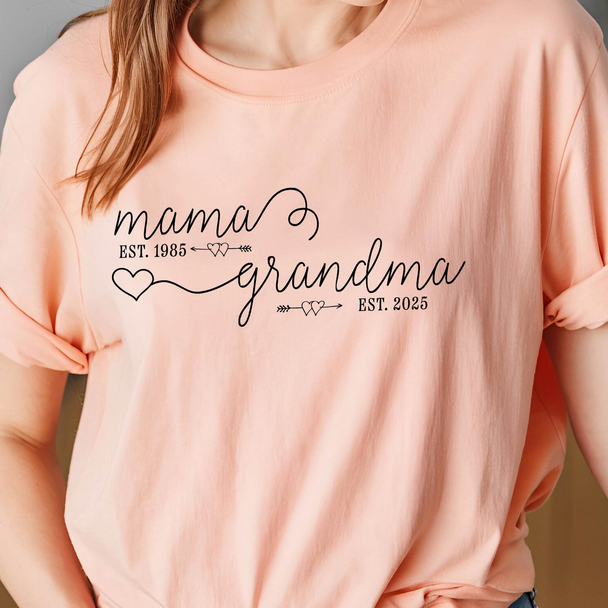 May include: A pink t-shirt with the text "mama est. 1985" and "grandma est. 2025" in black cursive font. The text is connected by a line with two small hearts and arrows.