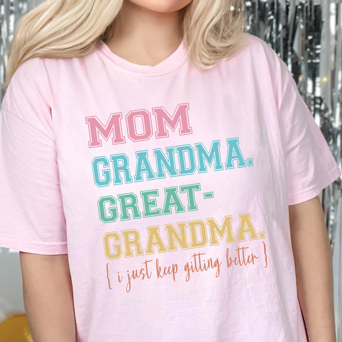 May include: Pink t-shirt with the text "MOM GRANDMA. GREAT-GRANDMA. [i just keep getting better]" in rainbow colors.