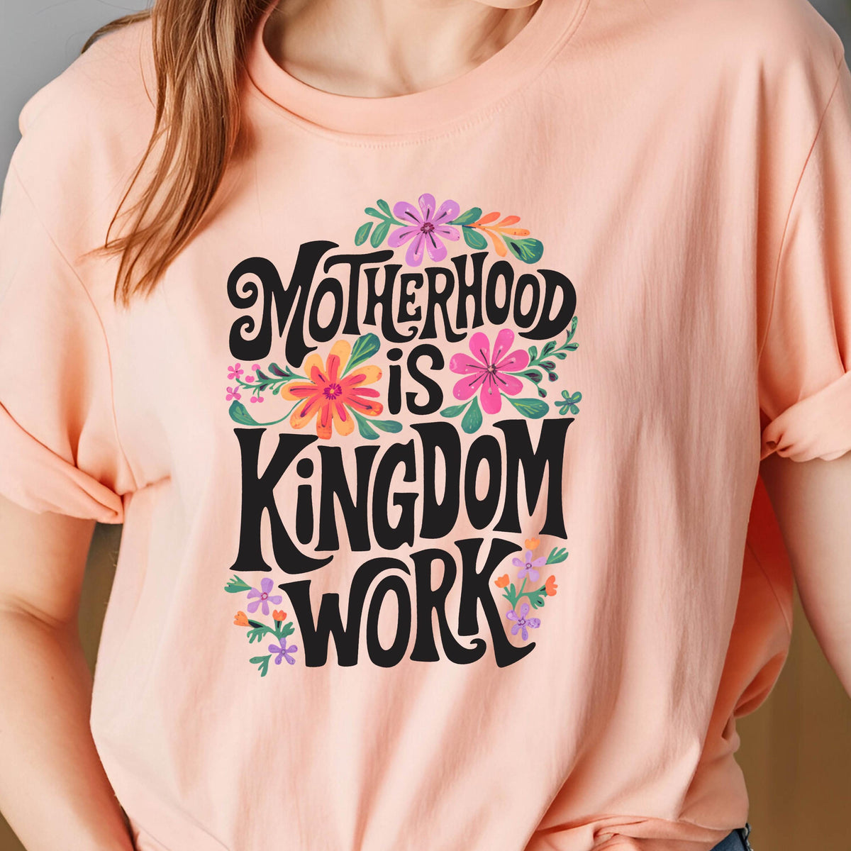 May include: A peach-colored t-shirt with a floral design and the text "Motherhood is Kingdom Work" in black lettering.