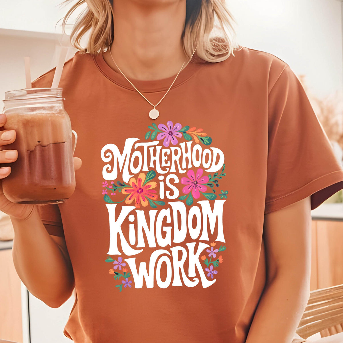 May include: A brown t-shirt with a white graphic that says "Motherhood is Kingdom Work" surrounded by colorful flowers.