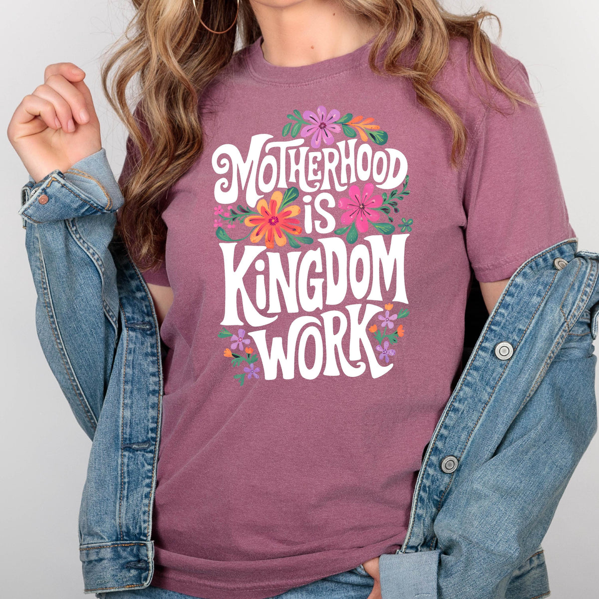 May include: A mauve t-shirt with a white graphic that reads "Motherhood is Kingdom Work" surrounded by colorful flowers.
