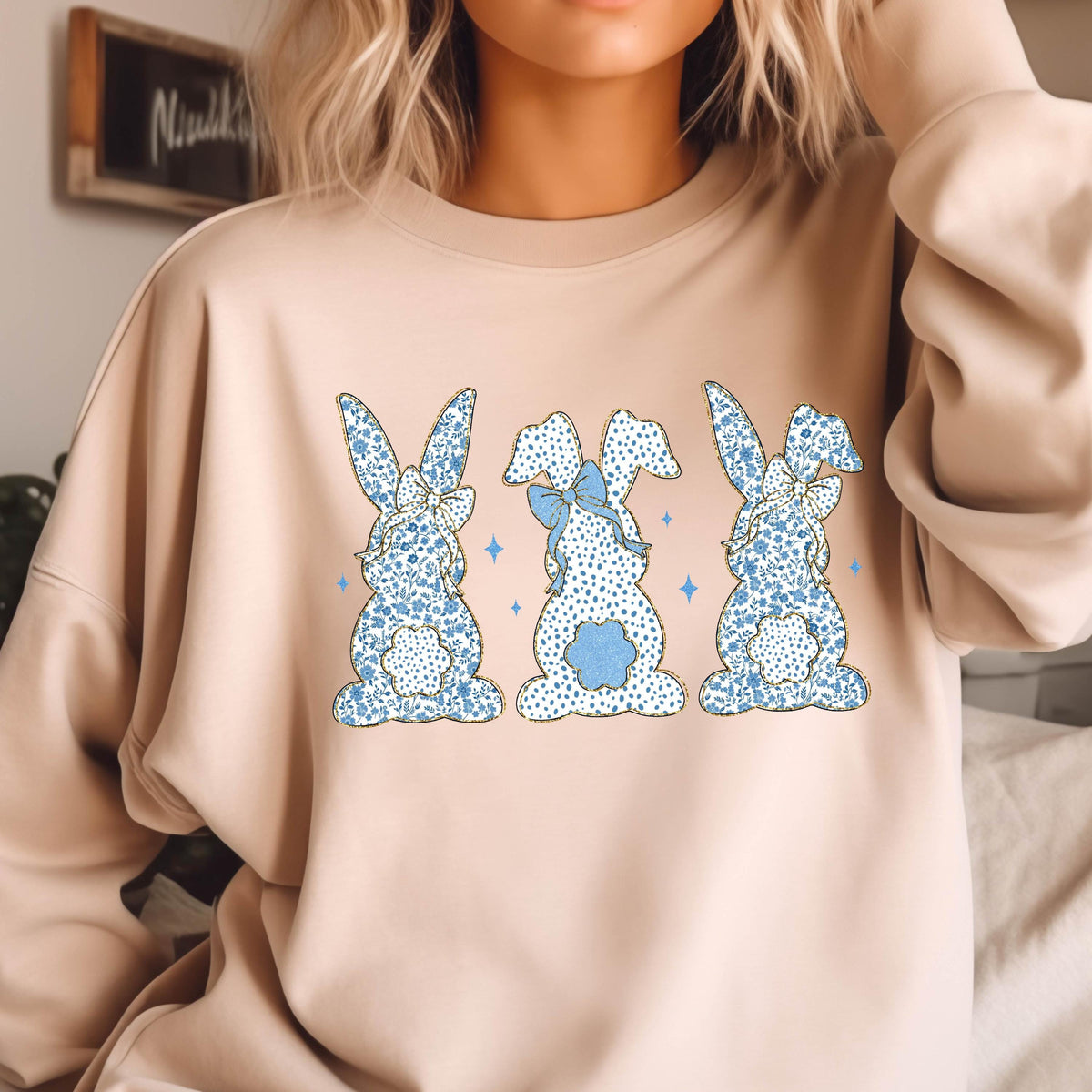 May include: A light beige sweatshirt with a graphic of three blue and white patterned bunnies with blue bows. The bunnies are facing the viewer and are arranged in a row.