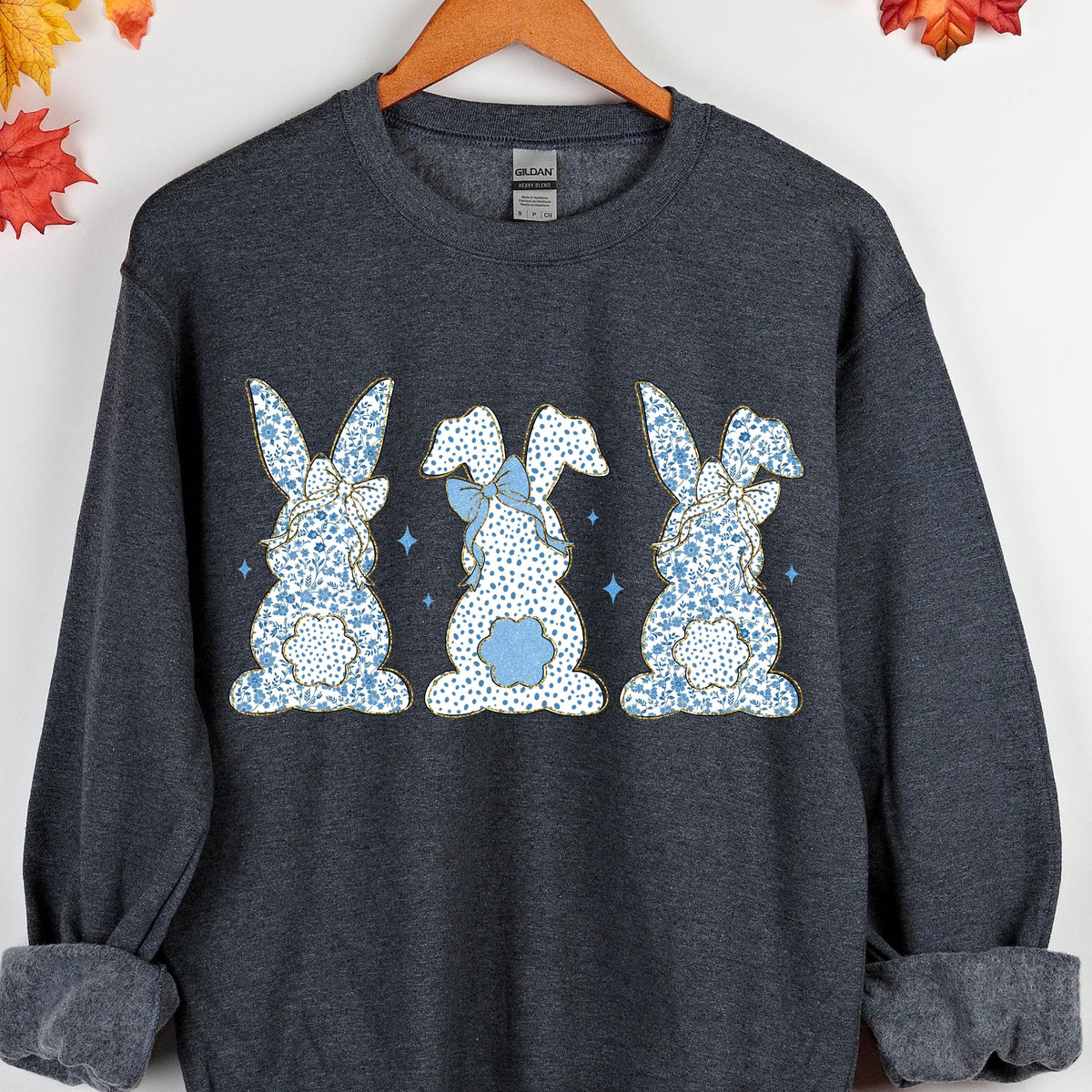 May include: A dark gray sweatshirt with a design of three blue and white bunnies with bows. The bunnies are facing forward and have a floral pattern.