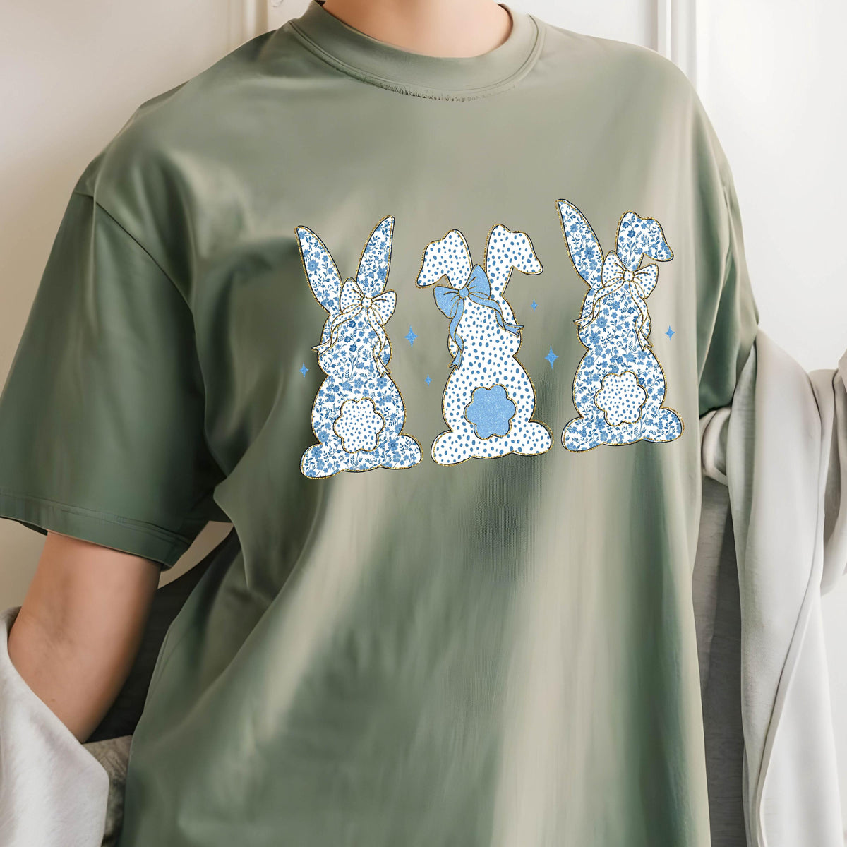 May include: A green t-shirt with a print of three blue bunnies with white polka dots and blue bows. The bunnies are facing forward and have their tails in the air.