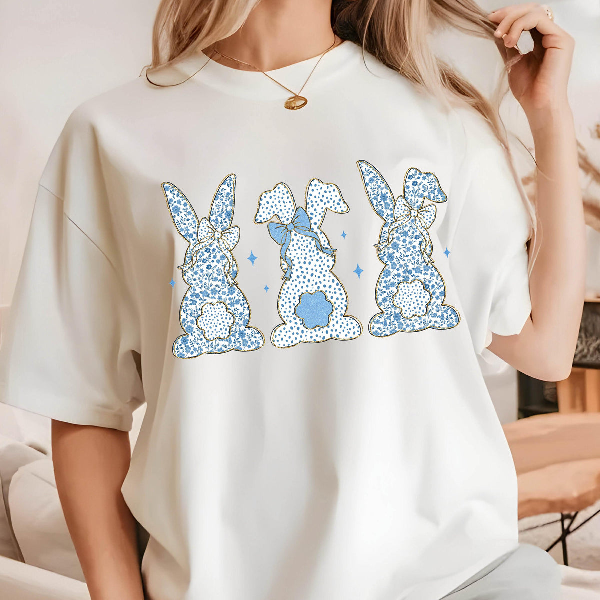 May include: White t-shirt with a graphic of three blue bunnies with gold outlines and blue bows. The bunnies are facing forward and have a floral pattern.