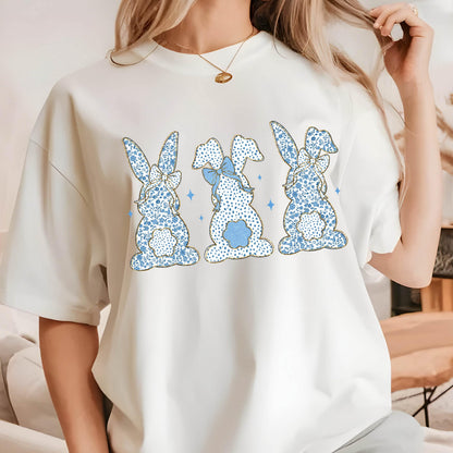 May include: White t-shirt with a graphic of three blue bunnies with gold outlines and blue bows. The bunnies are facing forward and have a floral pattern.