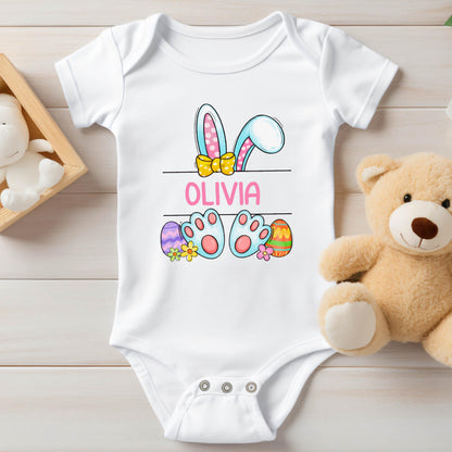 May include: A white short-sleeved baby bodysuit featuring a graphic of blue and pink bunny ears with a yellow bow, the name OLIVIA in pink text, and blue bunny feet surrounded by colorful Easter eggs and small flowers on the front center.