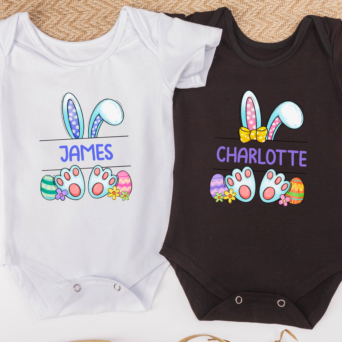 May include: Two white and black baby onesies with bunny ears and Easter eggs. The white onesie has the name "JAMES" printed on it. The black onesie has the name "CHARLOTTE" printed on it.