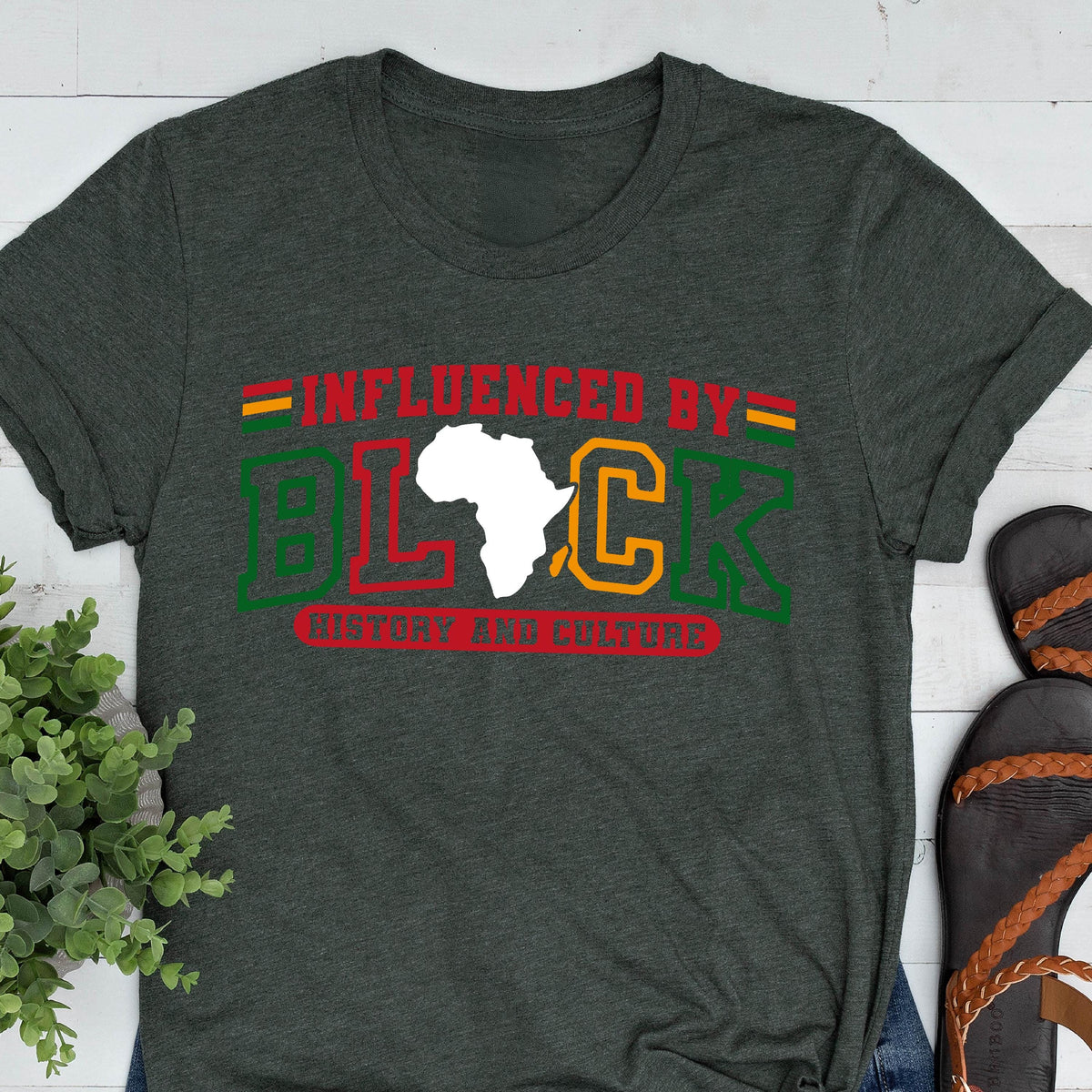 May include: A dark charcoal gray crew neck t-shirt featuring the text INFLUENCED BY BLACK HISTORY AND CULTURE in red, green, and yellow. The letter A in BLACK is replaced by a white silhouette of the African continent. Horizontal lines accent the text.