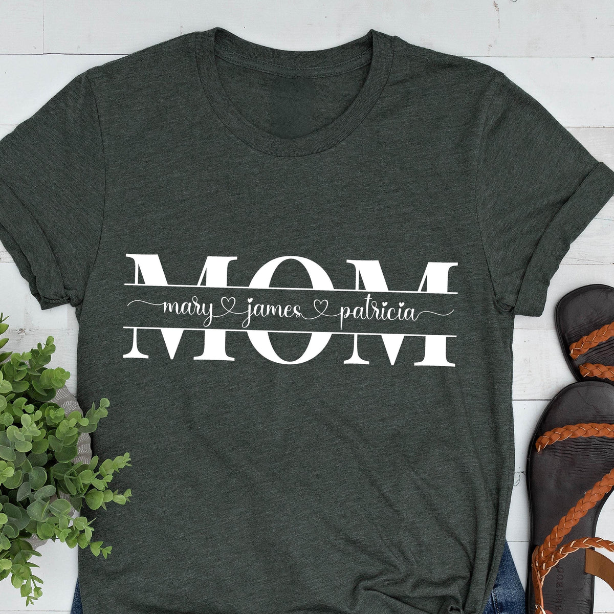 May include: A dark green t-shirt with the word "MOM" in white letters. The word "MOM" is separated by a line with the names "mary", "james", and "patricia" written in cursive with hearts between each name.
