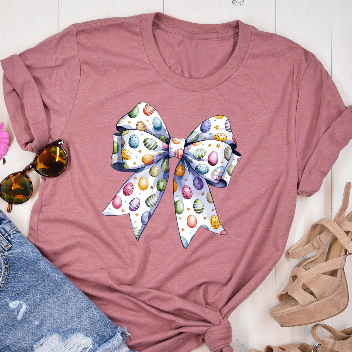 May include: A mauve t-shirt with a large white bow printed on the front. The bow is decorated with colorful Easter eggs and gold stars.