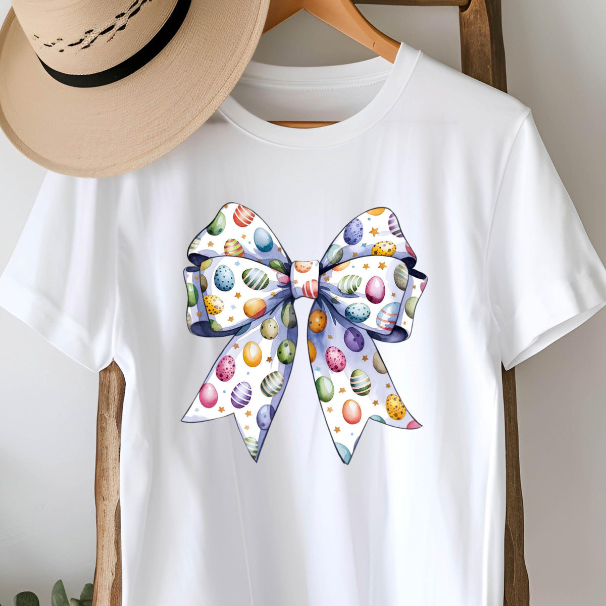 May include: White t-shirt with a large blue bow printed on the front. The bow is decorated with colorful Easter eggs.