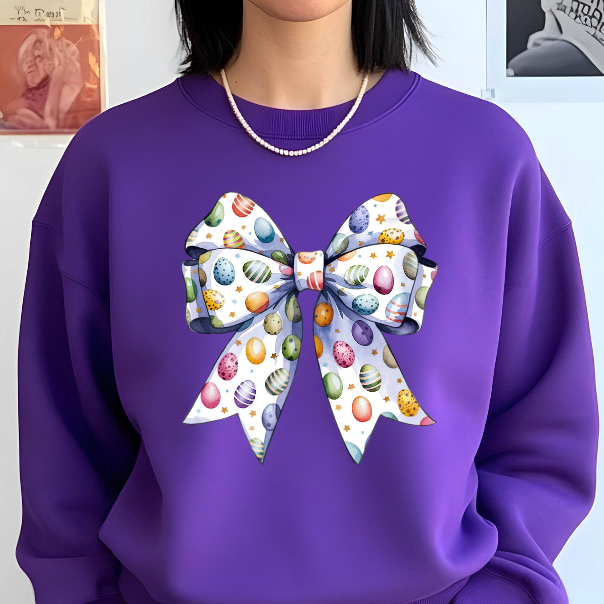 May include: Purple sweatshirt with a white bow decorated with colorful Easter eggs. The bow has a white background with gold accents.