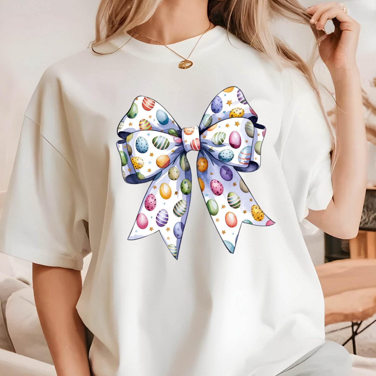 May include: White t-shirt featuring a large, colorful bow design. The bow is decorated with Easter eggs in various colors and patterns, with a light blue ribbon and gold stars. The shirt is a casual, everyday wear item.