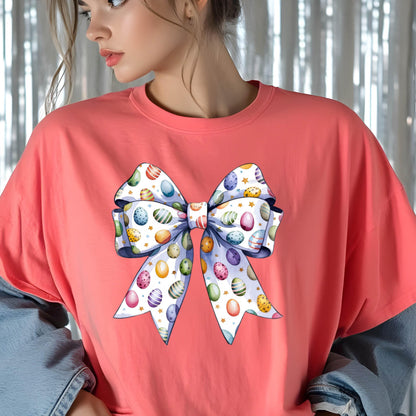 May include: Coral-colored t-shirt featuring a large bow design. The bow is decorated with colorful Easter eggs in various patterns and colors, including blue, yellow, and pink. The bow has a white background with gold stars.