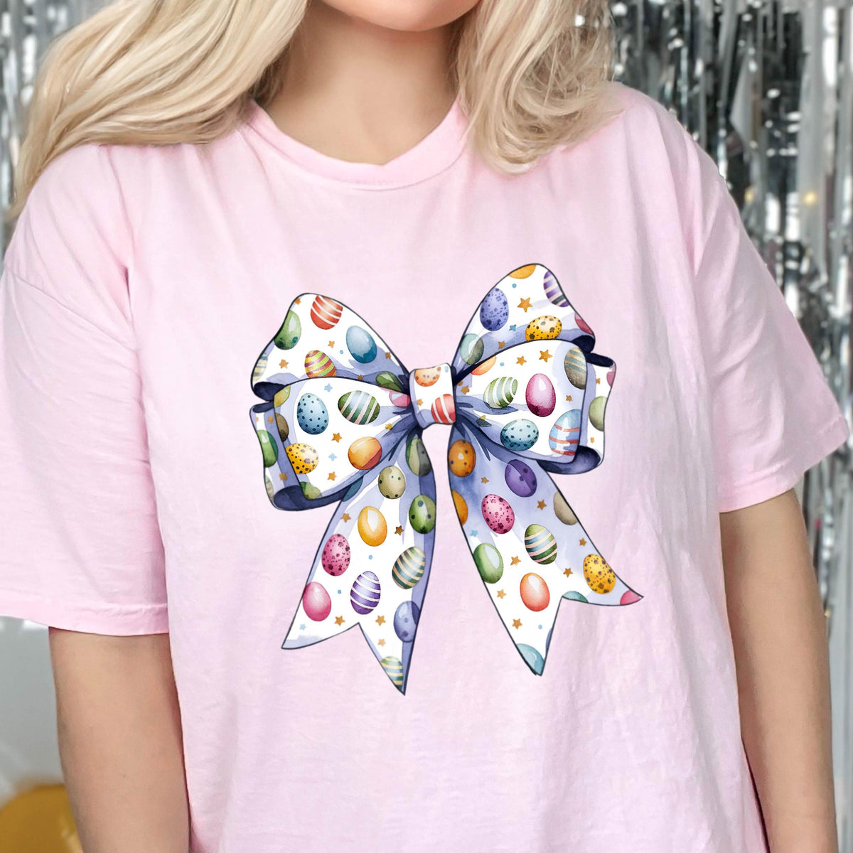 May include: A light pink t-shirt featuring a large, colorful bow design. The bow is decorated with Easter eggs in various colors, including blue, purple, yellow, and green, set against a white background with gold stars. The bow's edges are outlined in a soft blue.
