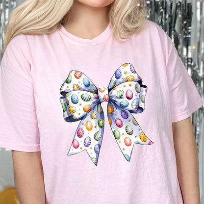 May include: A light pink t-shirt featuring a large, colorful bow design. The bow is decorated with Easter eggs in various colors, including blue, purple, yellow, and green, set against a white background with gold stars. The bow's edges are outlined in a soft blue.