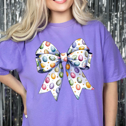May include: A purple t-shirt featuring a large bow design. The bow is decorated with colorful Easter eggs and small gold stars on a white background. The shirt has short sleeves.