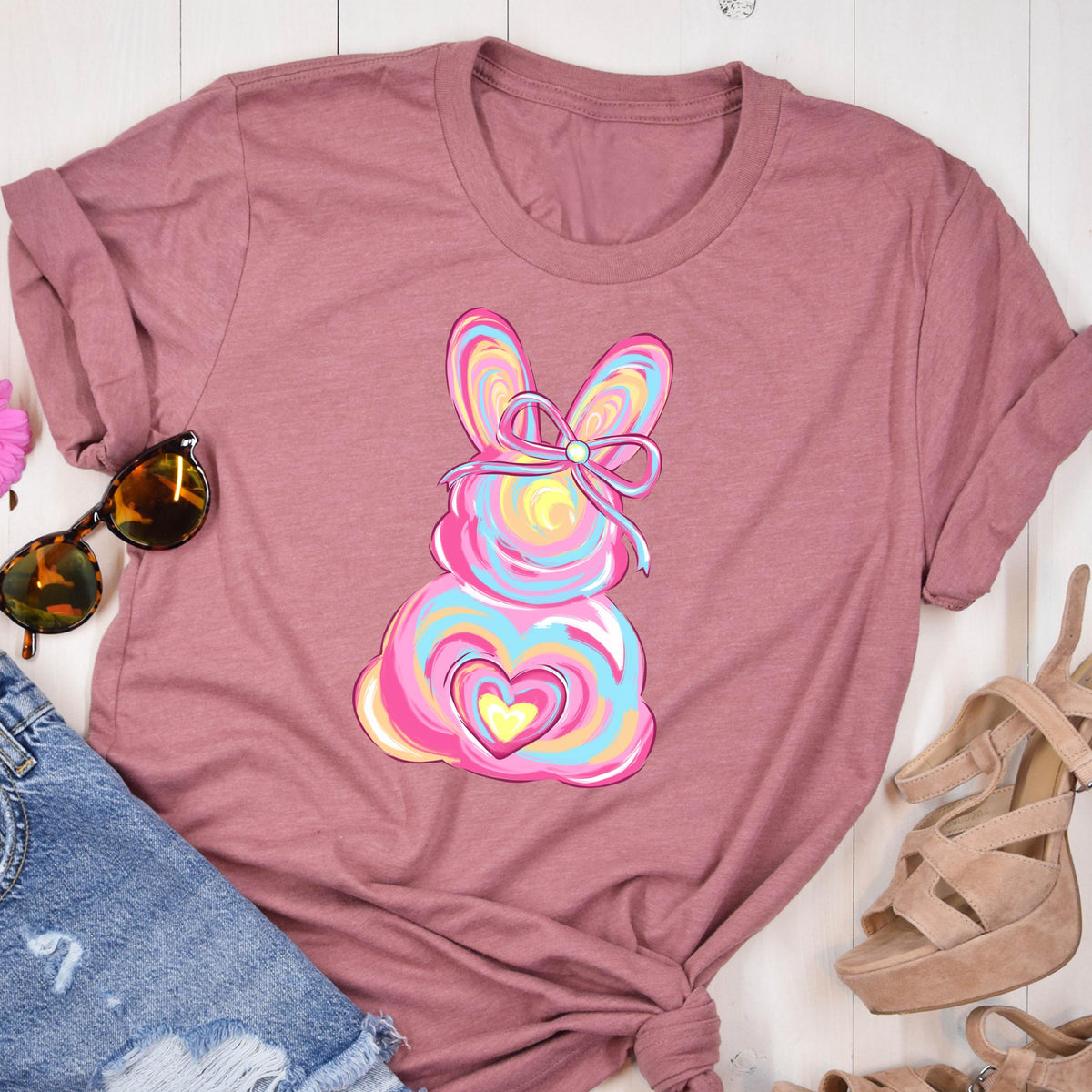 May include: A pink t-shirt with a colorful Easter bunny design. The bunny has a pink, blue, and yellow swirl pattern and a pink bow with a white pearl in the center. The bunny is holding a pink heart with a yellow heart inside.