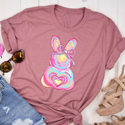 May include: A pink t-shirt with a colorful Easter bunny design. The bunny has a pink, blue, and yellow swirl pattern and a pink bow with a white pearl in the center. The bunny is holding a pink heart with a yellow heart inside.