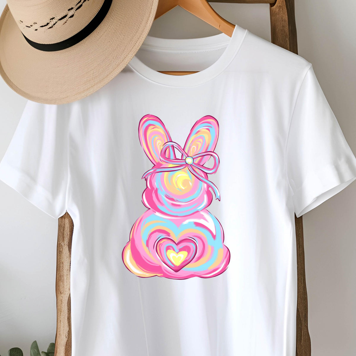 May include: White t-shirt with a pink, blue, and yellow swirl design of an Easter bunny with a pink bow. The bunny has a heart in the center of its body.