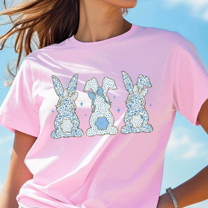 May include: Pink t-shirt with a graphic of three blue and white patterned bunnies with gold outlines. The bunnies are facing forward and have blue bows.