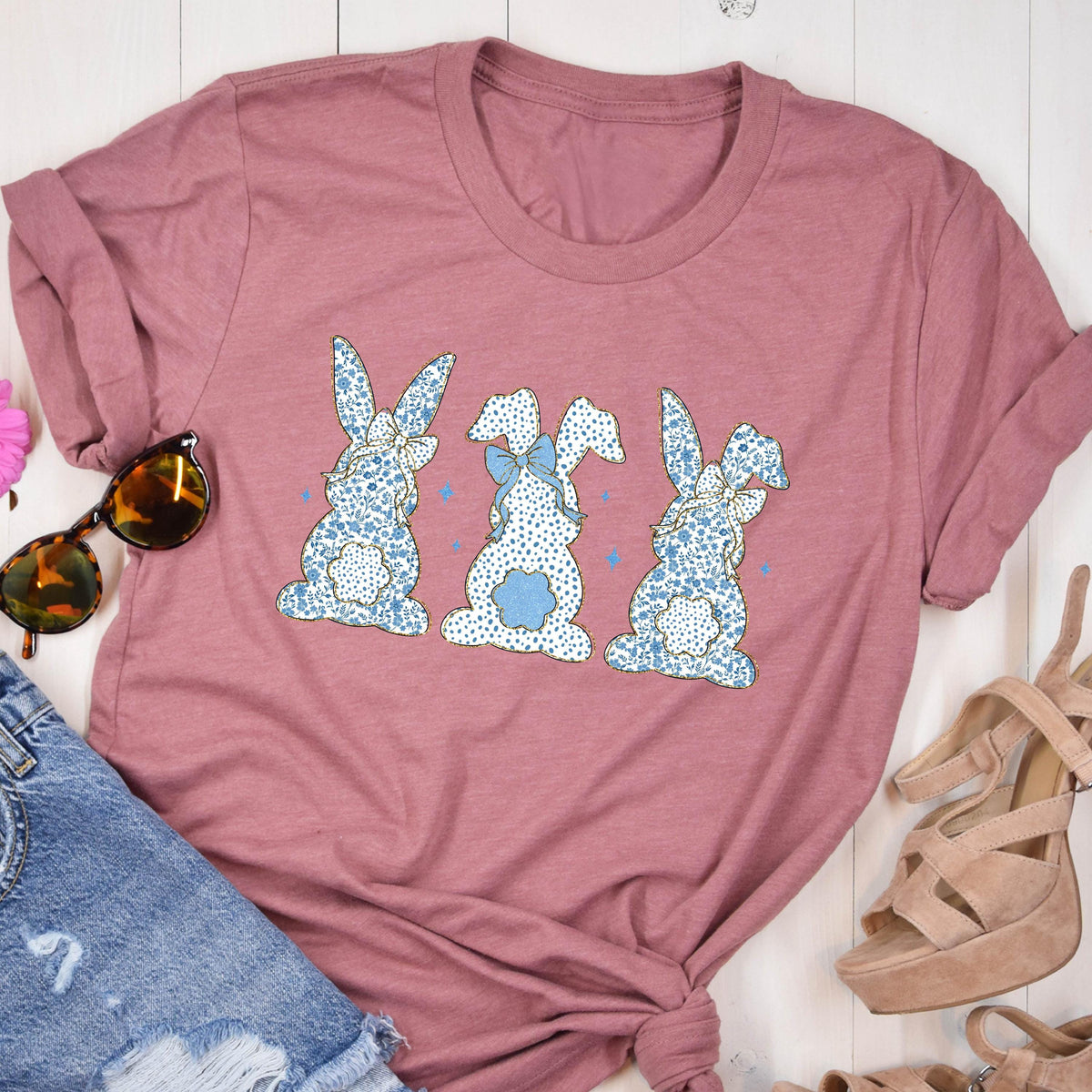 May include: A mauve colored t-shirt with a graphic of three blue and white patterned bunnies with bows. The bunnies are facing away from the viewer.