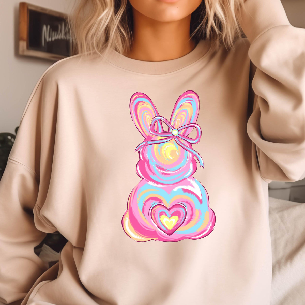 May include: A light brown sweatshirt with a pink, blue, and yellow swirl design of an Easter bunny with a pink bow. The bunny has a heart shape in the center.