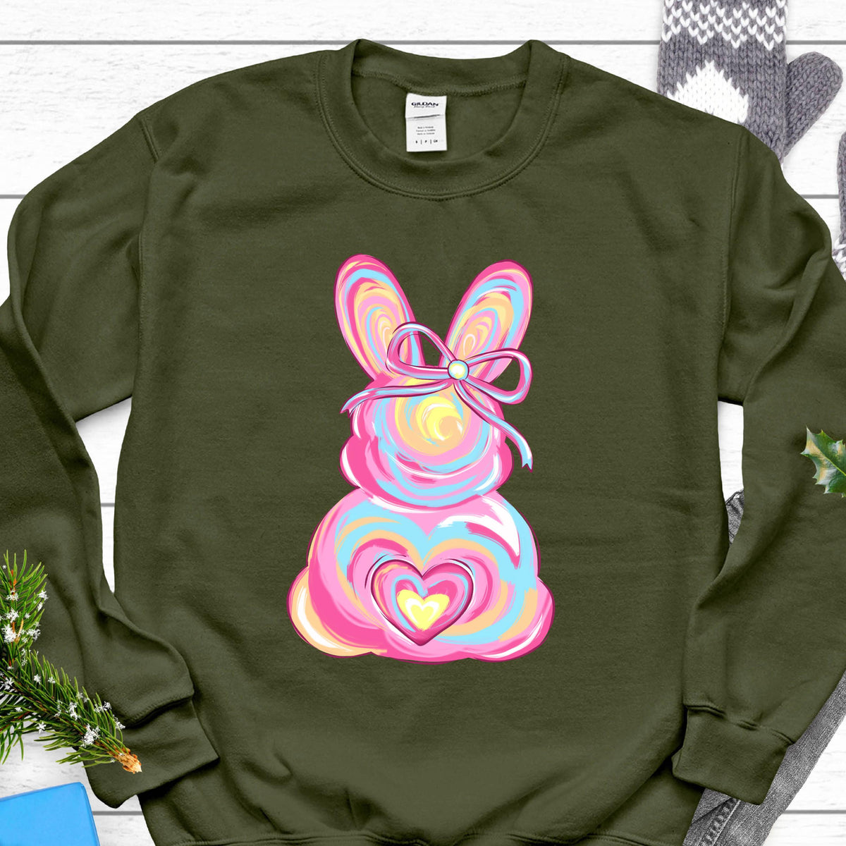 May include: A green sweatshirt with a pink, blue, and yellow bunny rabbit design. The bunny has a pink bow and a heart in the center of its body.