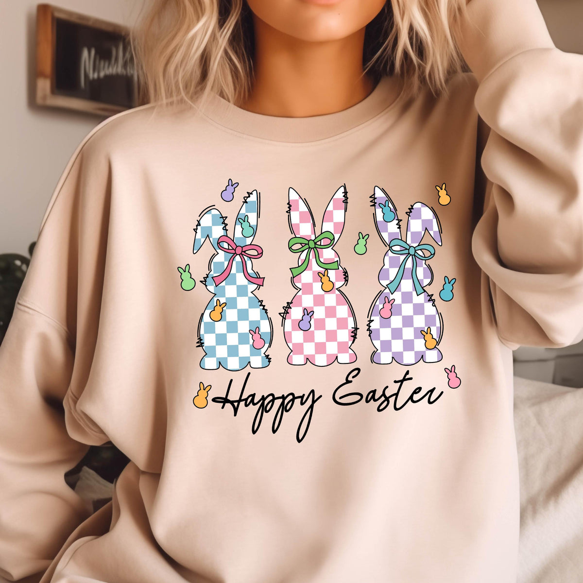 May include: A light beige sweatshirt with a graphic of three Easter bunnies in a row. The bunnies are wearing checkered dresses with bows and are surrounded by pastel-colored jelly beans. The text "Happy Easter" is printed below the bunnies.